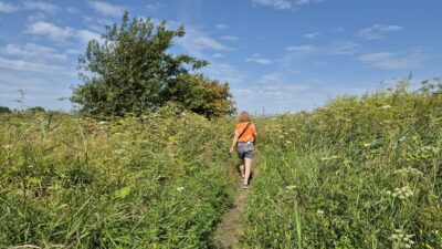 Dame met korte broek en oranje shirt loopt buiten tussen de planten. Blauwe lucht met kleine wolken.